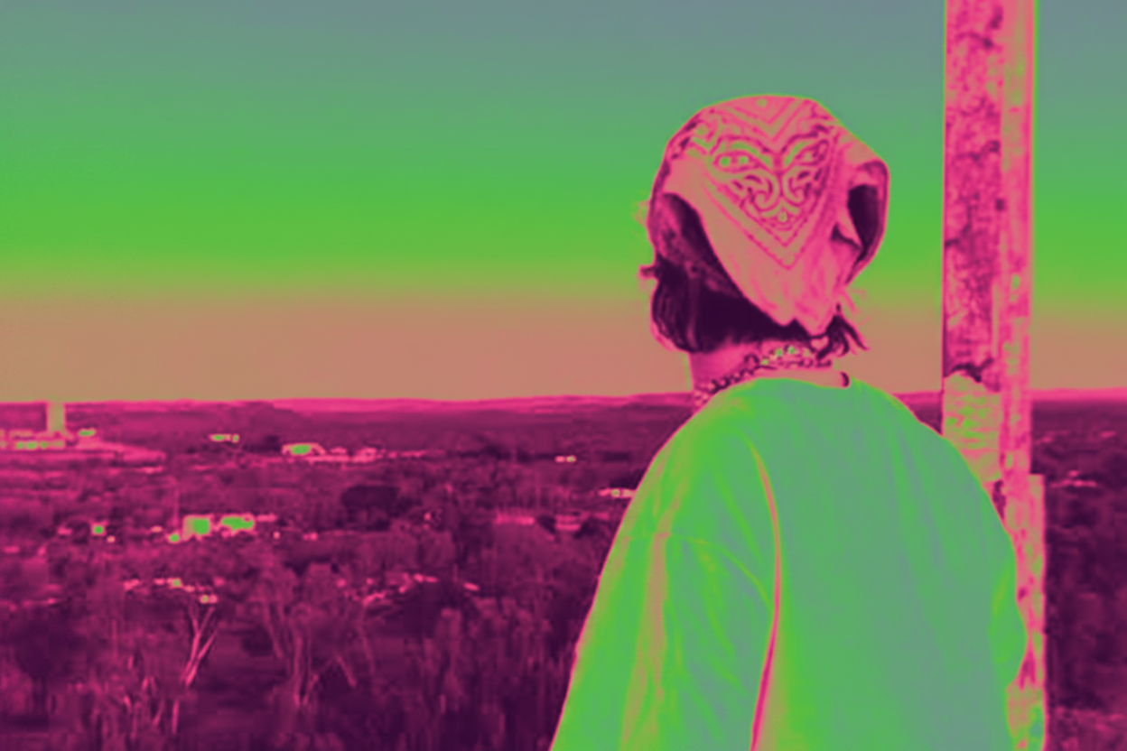 A person in a bright neon green outfit sits on a weathered wooden structure overlooking a wide landscape, with a strong pink and green color-grading effect across the entire scene.